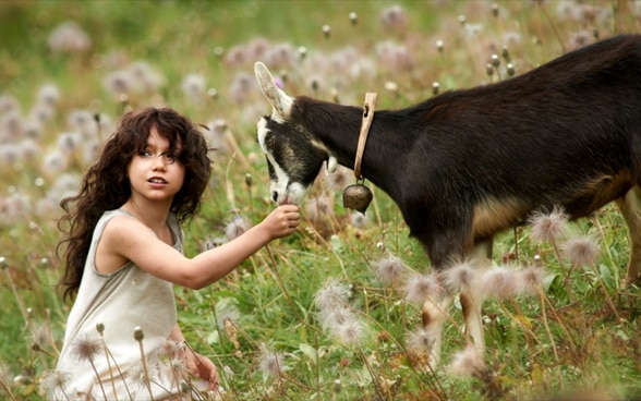 Una scena di un film: Heidi in un pascolo di montagna fiorito insieme a una capra. 