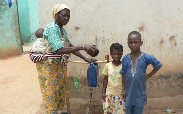A woman with an infant on her back shows two children how to fill the makeshift handwashing device with water in front of a hut.