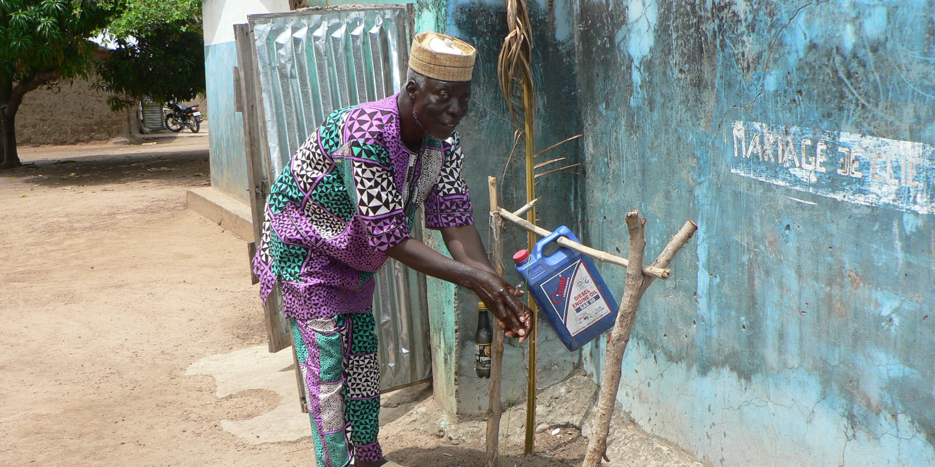 In Benin, a man stands in front of a hut and washes his hands thoroughly at a makeshift handwashing station.