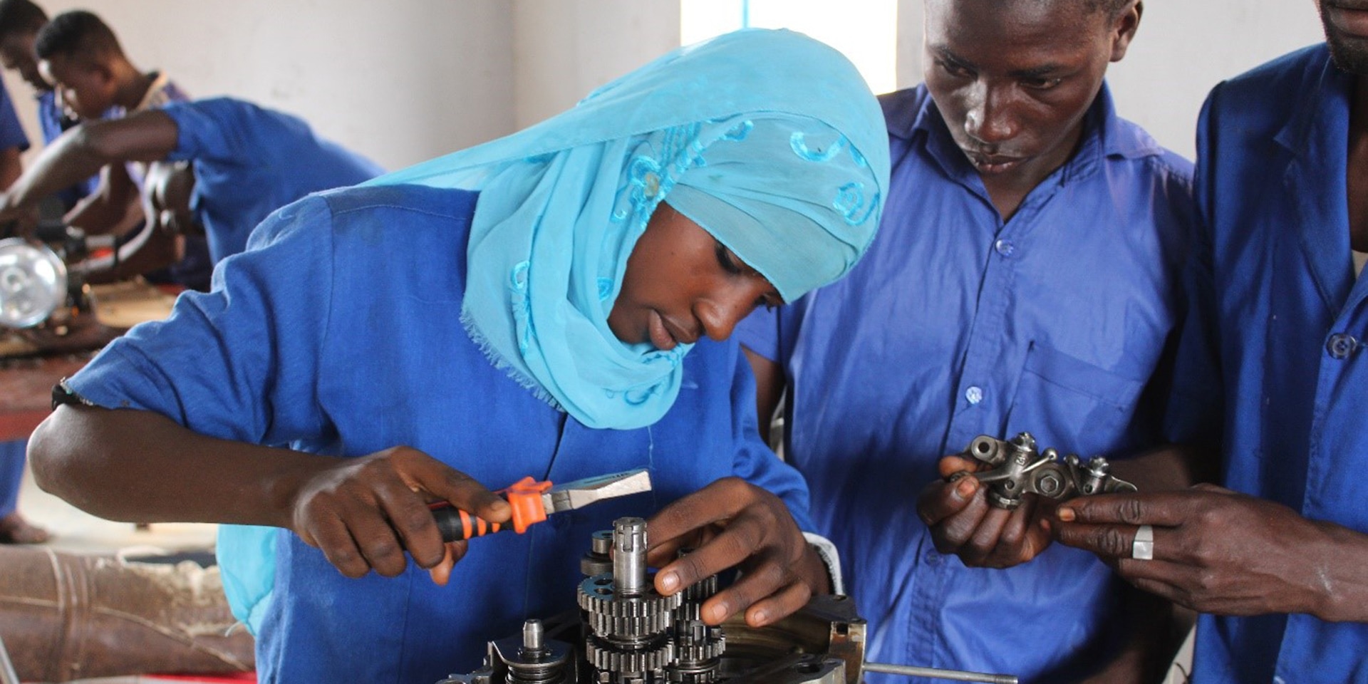 In a training workshop, a young woman with a head covering works on a mechanical component, observed by two male apprentices.