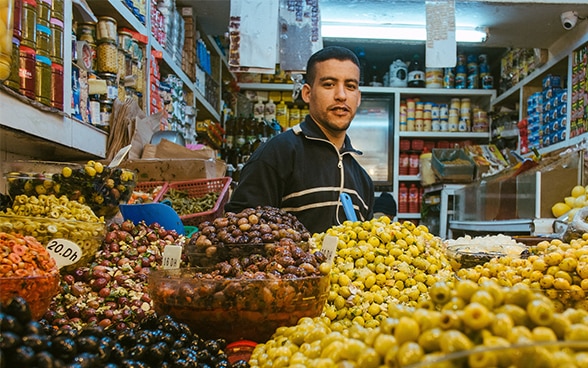 Im Bild sieht man einen Mann in einem kleinen Laden vor einer grossen Auslage mit Oliven.