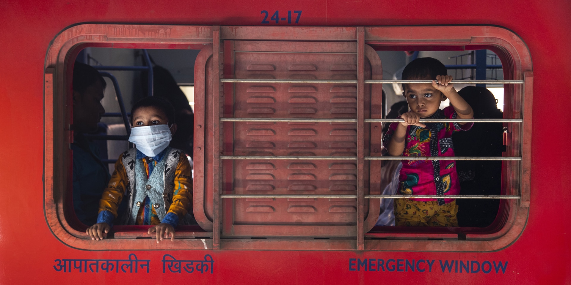 Two children look out of the sliding window of a booth – one with a mask, one without.