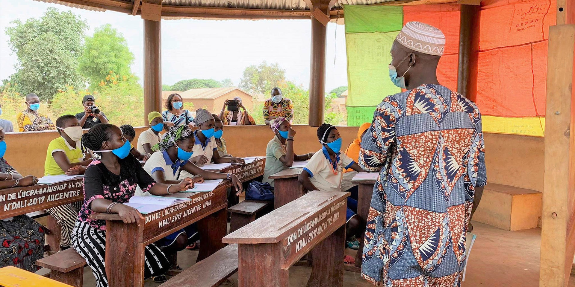  Schülerinnen und Schüler in einer Schule in Benin sitzen an Holztischen und werden von einem Lehrer unterrichtet. Das Dach des Klassenzimmers wird von Holzpfählen gestützt. 