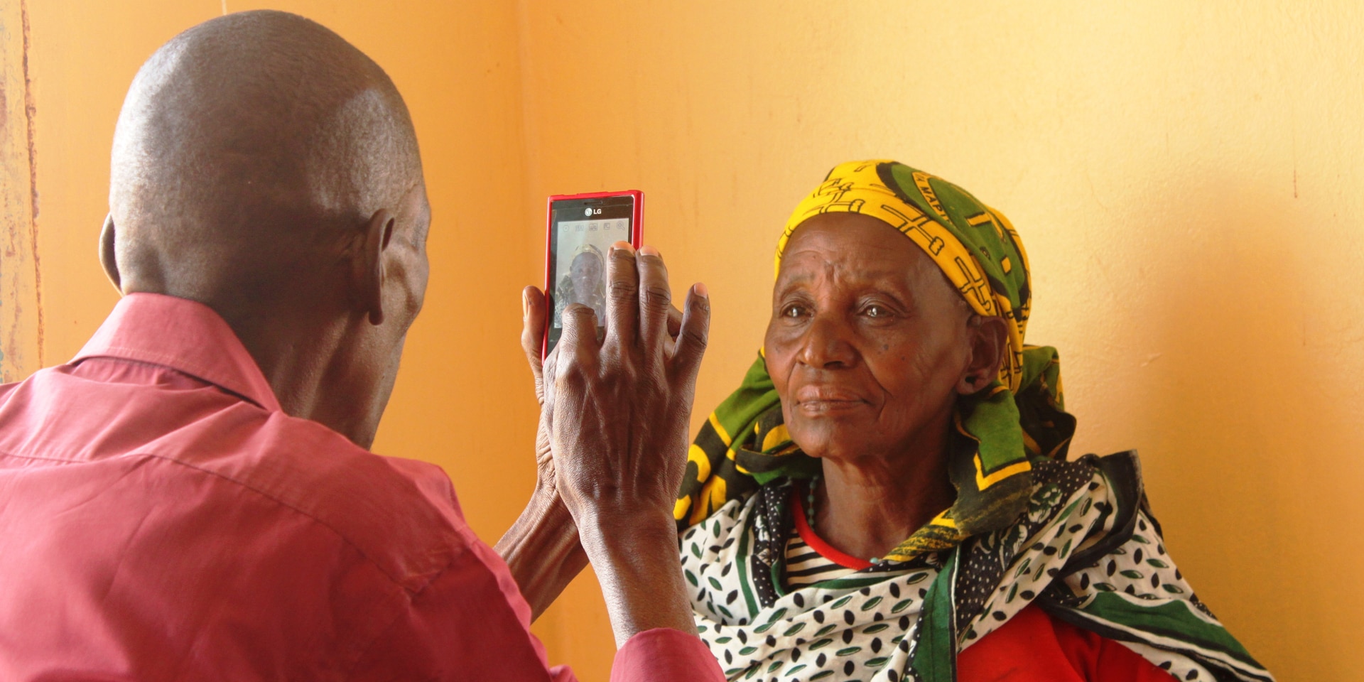 Un homme prend une femme en photo avec son téléphone portable. La femme se tient devant un mur orange. 