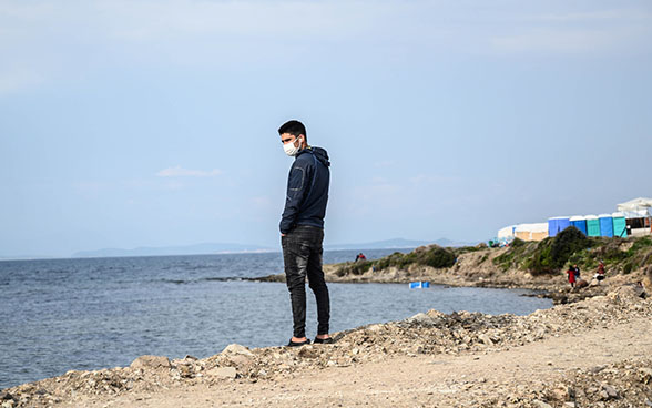 A migrant stands by the sea in the Kara Tepe refugee camp on the Greek island of Lesbos, 29 March 2021. The upper-right of the image shows tents along the coast, with women dressed in red standing by the sea.