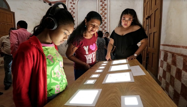 Una niña con auriculares mira las imágenes en una mesa. Junto a ella hay dos mujeres.