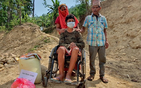 Un homme et une femme poussent un jeune homme en fauteuil roulant à travers un terrain vague au Népal.
