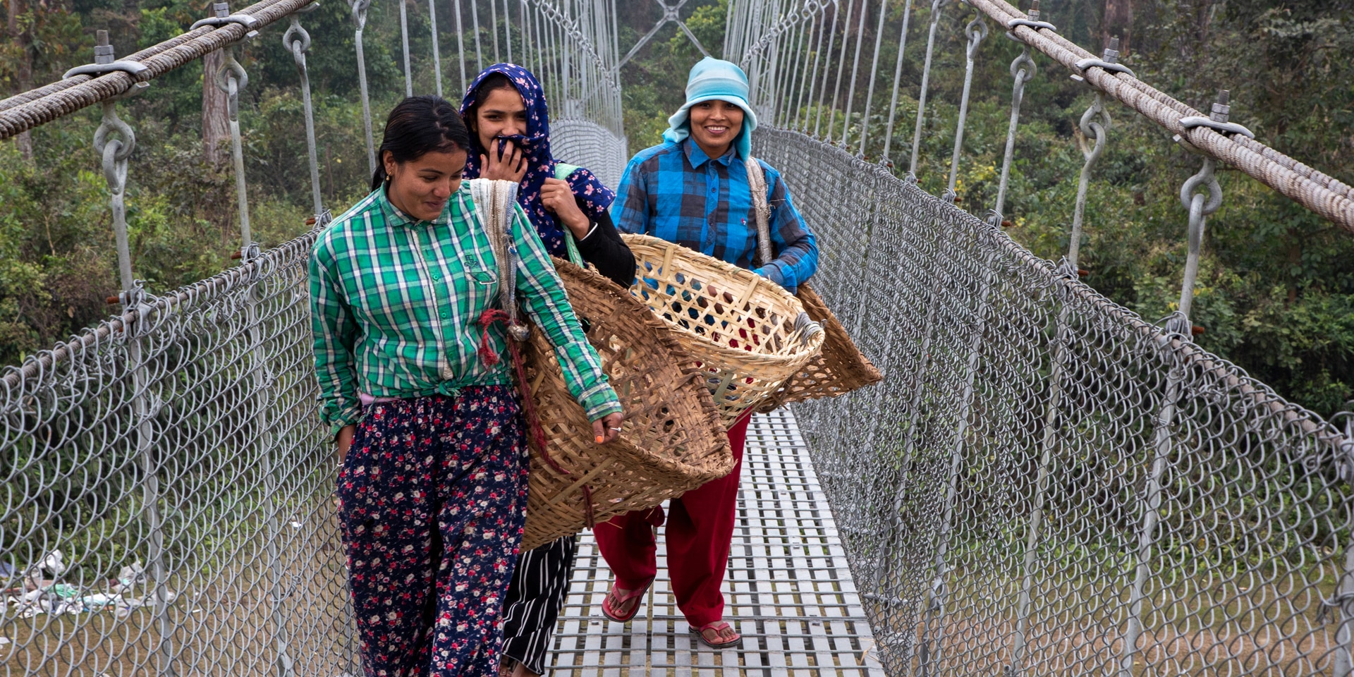 Tres mujeres cruzan un puente nepalí sonriendo.