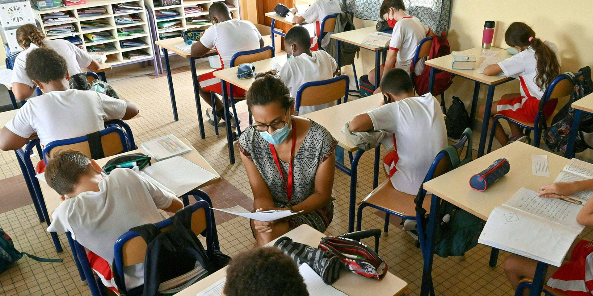 Kinder sitzen zwischen Schulbänken. Die Lehrerin hilft ihnen bei ihren Schulaufgaben.