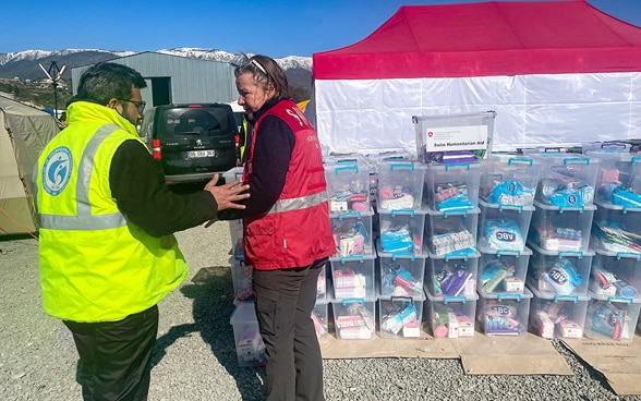 Una collaboratrice del Corpo svizzero di aiuto umanitario con un gilet rosso e un uomo con un gilet giallo sono in piedi davanti a scatole contenenti kit per l'igiene personale.