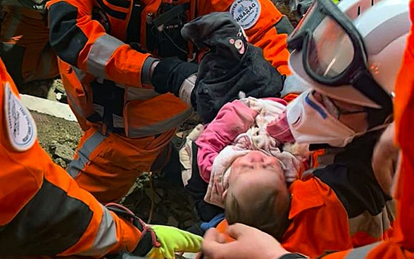 Un bambino giace tra le braccia di una donna con casco e vestiti arancioni, intorno a lei altri due membri della catena di soccorso.