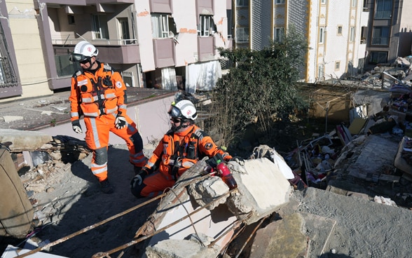 Due uomini vestiti di arancione si trovano sulle macerie di una casa crollata.