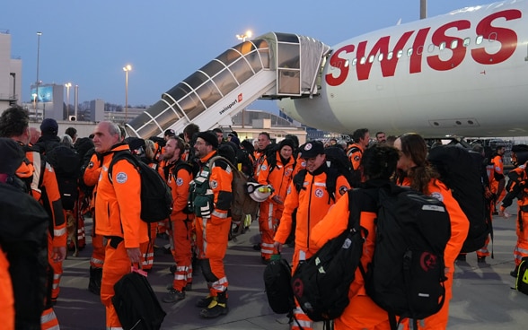 Numerosi membri della catena di soccorso in tenuta arancione si trovano sul campo di volo. Sullo sfondo si vede un aereo con una scala agganciata.
