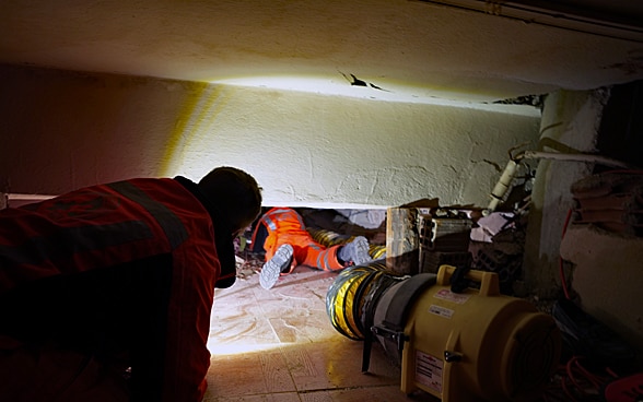 Un membre de la Chaîne suisse de sauvetage rampe sous les décombres d'une maison. Un projecteur l'éclaire.