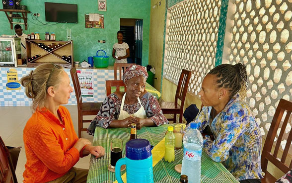 La directora de la COSUDE, Patricia Danzi, y una colega de la oficina de cooperación suiza en Benín sentadas a una mesa conversando con la dueña de un restaurante.