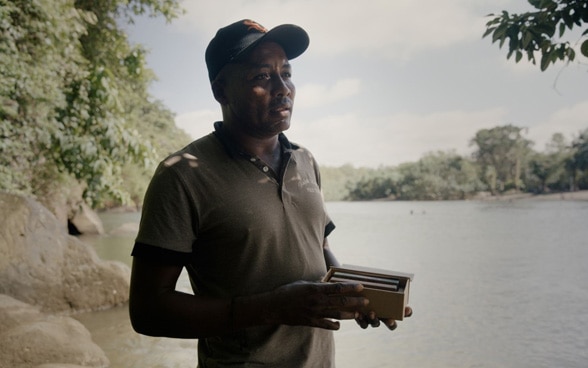 Un homme tient une boîte de chocolat, debout au bord d’une rivière.