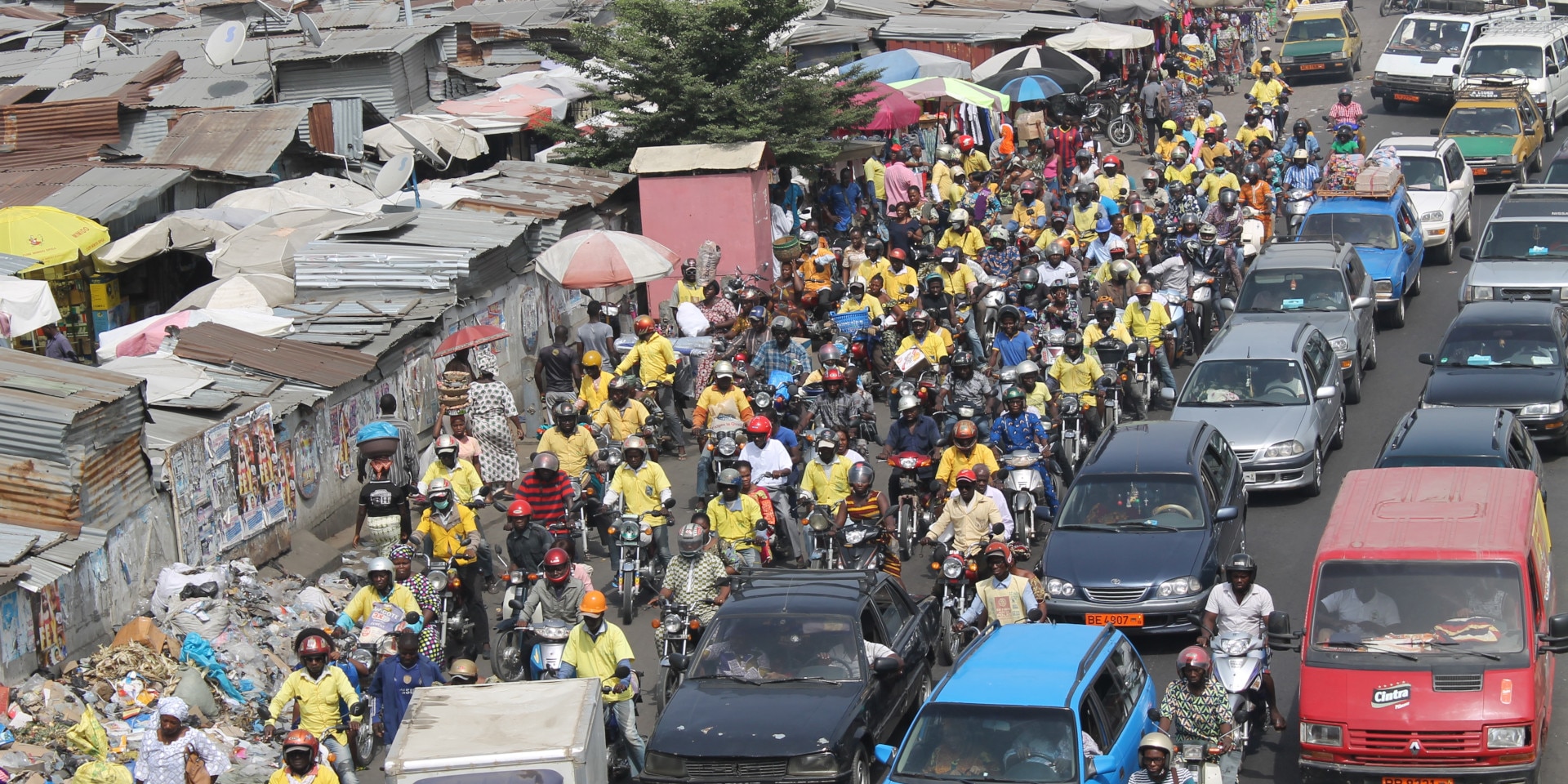 Des voitures et des motos circulent sur une route du Bénin. Des logements de fortune ont été construits le long de la route.