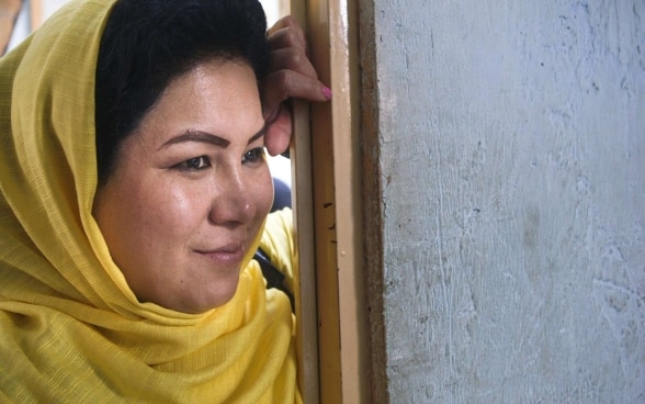 Eine Afghanische Frau mit hellgelbem traditionellen Kopftuch leht lächelnd an einem Türrahmen.