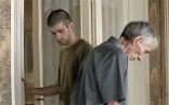 A middle-aged and a young man stand shoulder to shoulder, looking at the floor in opposite directions.  