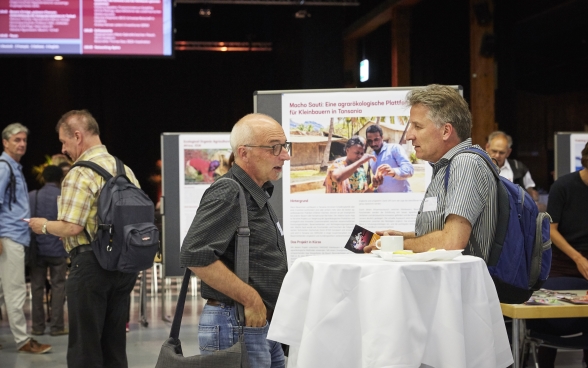 Two participants by a standing table at the Bernexpo Festhalle. Behind them is the exhibition from the different partner organisations.