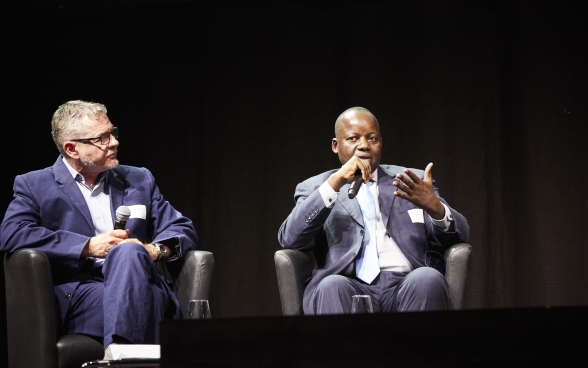 Mansour N’Diaye sitting on stage and speaking into a microphone. Dominique Guenat sitting next to him and listening attentively.
