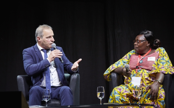 Pierre-André Page sitting on stage and speaking into a microphone. Mariane Nguerassem sitting next to him listening attentively.