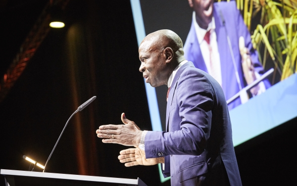 Gilbert F. Houngbo standing behind a lectern on stage. The screen on which the speech is being transmitted can be seen in the background.
