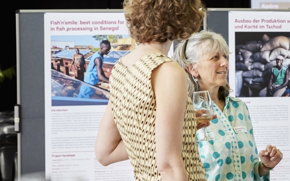 Zwei Teilnehmerinnen stehen vor einem Poster der Ausstellung der Partnerorganisationen und unterhalten sich.