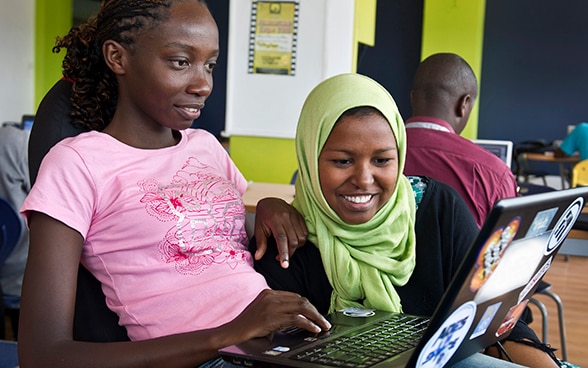 Two smiling women sitting next to each other surf the internet on a laptop while a man behind them is using his laptop. 