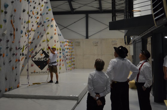 Three Swiss musicians in traditional attire watch two men negotiating an indoor climbing wall.