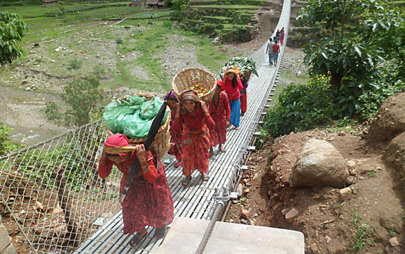 Varias mujeres cargadas con cestas atraviesan un puente colgante.