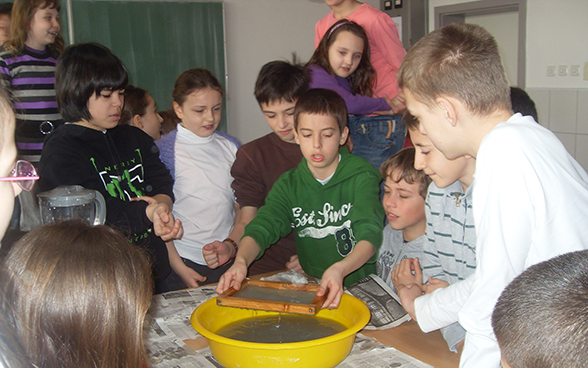 Jungen und Mädchen schauen einem Mitschüler zu, der ein Wasserexperiment vorführt.