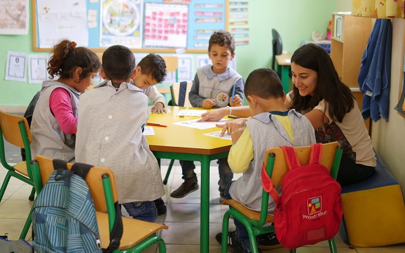 Inside a classroom where a woman teacher is sitting in front of a group of children showing them a book. 