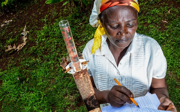 Una giovane contadina keniota di nome Mercy Wambui sta in piedi sull'erba. Misura l'acqua piovana e inserisce i dati nella sua lista.