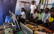 a man repairing a computer in his workshop, watched by six visitors.