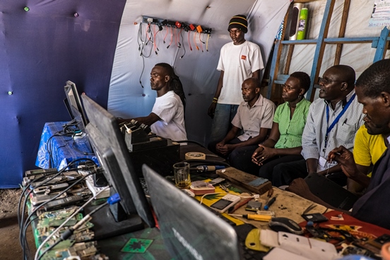 un homme répare un ordinateur dans son atelier, devant 6 visiteurs.