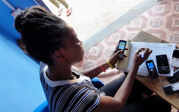 An African woman sits at a table on which several mobile phones are lying and transfers numbers into a table.