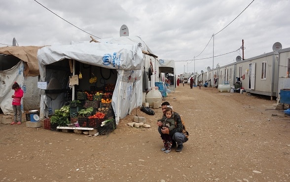 Un père et sa fille à côté d’un stand de fruits et de légumes dans un camp.