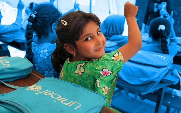 Dans une salle de classe, des petites filles lèvent la main pour répondre aux questions posées par la maîtresse; l’une d’elles se retourne pour sourire à l’objectif.