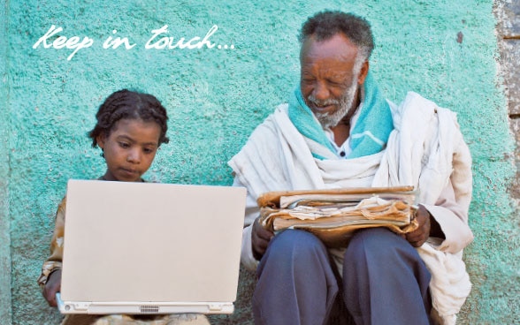 A young african girl holding a laptop and an old man holding newspapers, sitting next to each other.
