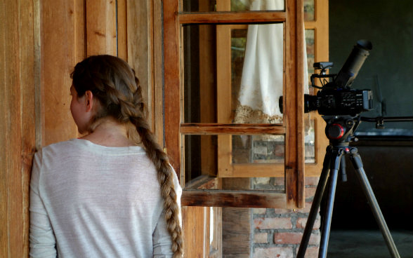 The photo shows Medea from the back with a film camera in front of a window to her right.