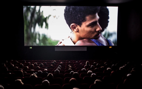 Blick in einen dunklen Kinosaal, auf der Leinwand umarmen sich zwei Männer.