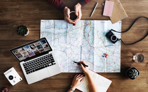 Due persone sedute a un tavolo stanno pianificando un viaggio munite di carta geografica, penna, agenda e laptop.