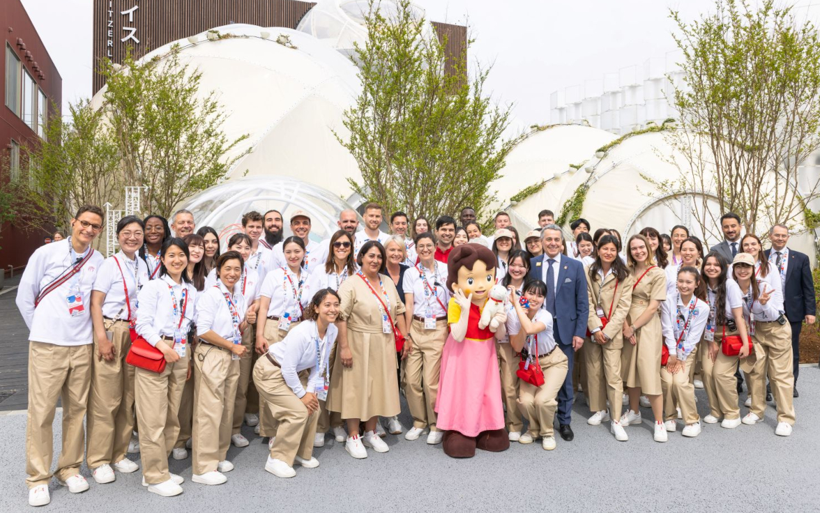 Un grande gruppo di persone in uniforme posa con una persona vestita da Heidi davanti a edifici a forma di cupola e ad alberi.