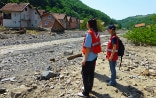 Due donne con giubbotto catarifrangente osservano una casa distrutta sulla riva di un torrente.