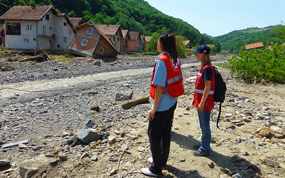 Due donne con giubbotto catarifrangente osservano una casa distrutta sulla riva di un torrente.