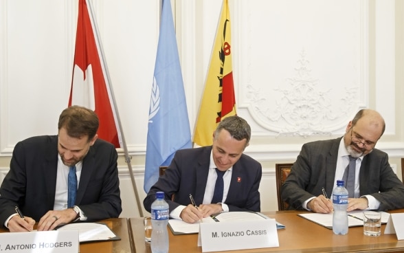 Bundesrat Cassis, Antonio Hodgers und Sami Kanaan sitzen an einem hölzernen Tisch und unterzeichnen die gemeinsame Erklärung. 