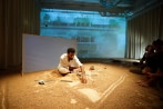 Un uomo seduto sul palcoscenico disegna sulla sabbia; il pubblico è seduto per terra, vicino all’artista. Dietro l’artista viene proiettato un film su uno schermo. 