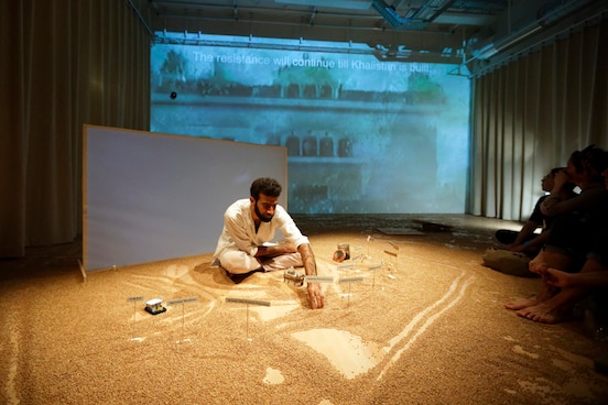 Ein Mann sitzt auf der Bühne und zeichnet im Sand, das Publikum sitzt ganz nah vom Künstler auf dem Boden. Hinter dem Künstler wird auf einer Leinwand ein Film gezeigt. 