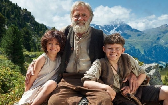 Alp-Öhi, the grandfather of the Heidi film adaption is sitting in front of mountain backdrop, holding his arms around Heidi and Peter.